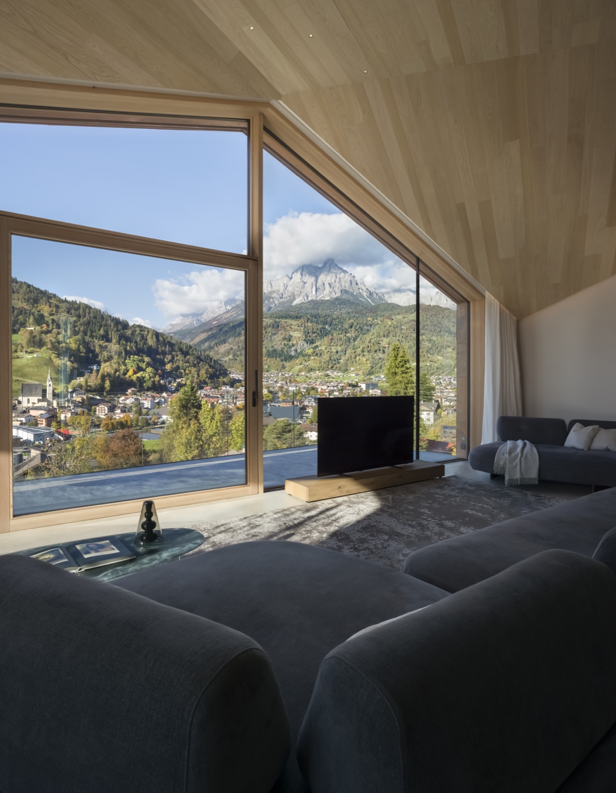 Il soggiorno di Casa LAGO Dolomiti con grandi vetrate aperte sul paesaggio alpino e sul borgo di Primiero
